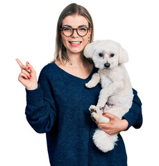 Young blonde woman hugging cute dog smiling happy pointing with hand and finger to the side