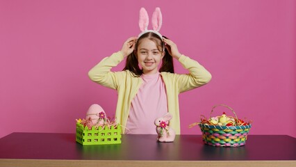 Obraz premium Smiling confident toddler placing bunny ears on her head and waving, saying hello against pink background. Cute joyful child creating decorations for easter sunday holiday event. Camera B.