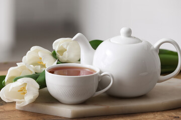 Board with hot brewed cup of tea, pot and tulips on table against blurred background