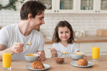 Obraz na płótnie Canvas Happy young caucasian father eating breakfast with his lovely little daughter in the kitchen, spending time together. Family holliday, celebration. Happy father`s day! I love you, dad!