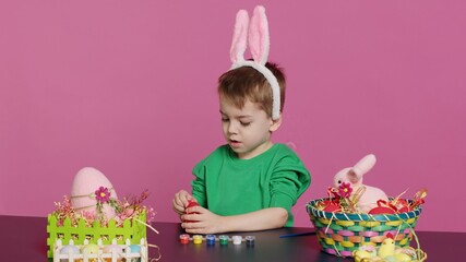Obraz premium Adorable little youngster creates handcrafted ornaments for Easter Sunday and painting eggs with stamps and brushes. Pleased young boy using art materials to color and decorate. Camera A.