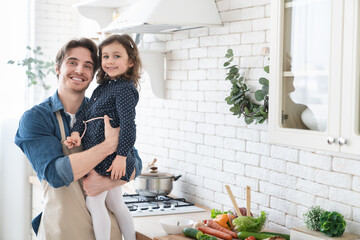 Happy young father enjoying cooking with his little daughter in the kitchen, preparing family dinner together, ready for celebrating father`s day. Parenthood and fatherhood