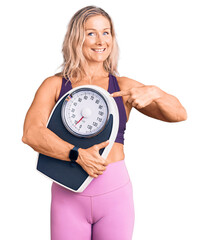 Middle age fit blonde woman wearing sports clothes holding weighing machine smiling happy pointing...