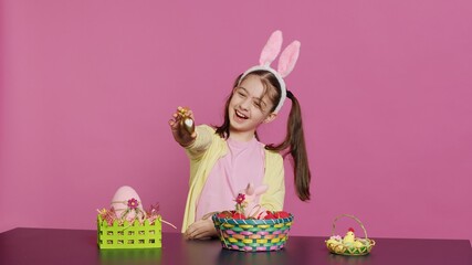 Obraz premium Excited young girl arranging painted eggs in a basket to prepare for easter holiday celebration, creating festive arrangements. Playful happy toddler with bunny ears, creative activity. Camera A.