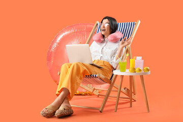 Young businesswoman with laptop and beach accessories ready for summer vacation on orange background