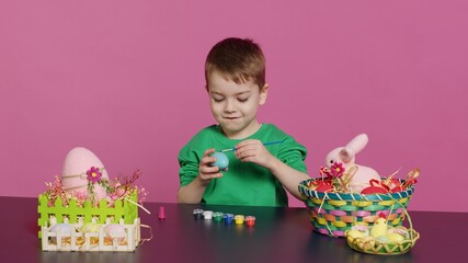 Obraz premium Little cheerful boy crafting handmade easter decorations by painting with watercolor and paintbrushes. Small young kid using art supplies coloring eggs for holiday, decorating activity. Camera A.
