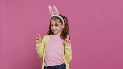 Lovely sweet girl posing in studio with fingers crossed and hoping to receive gifts for easter holiday celebration. Cheerful young child with bunny ears asking for good luck and fortune. Camera A.