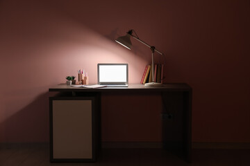 Table with modern laptop and glowing lamp near pink wall
