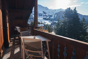 Balcony view of snowy mountains, wooden chalet with table and chairs. Overlooks alpine valley with buildings. Clear sky, alpine scenery in Europe or North America.