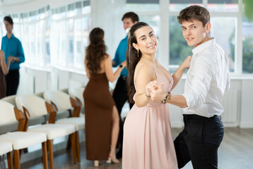 Stylish emotional hispanic guy in white shirt and black trousers enjoying impassioned merengue with young female partner wearing elegant dress in latin dance class. Amateur dancing concept.. - obrazy, fototapety, plakaty