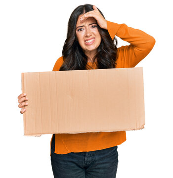 Beautiful brunette young woman holding cardboard blank empty banner stressed and frustrated with hand on head, surprised and angry face