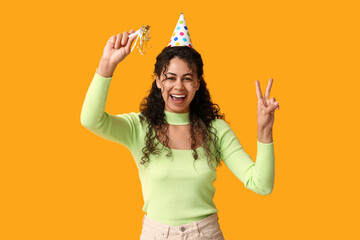 Beautiful young African-American woman in party hat with whistle showing victory gesture on yellow...
