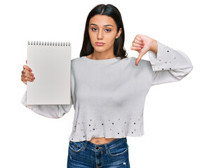 Young hispanic girl holding notebook with angry face, negative sign showing dislike with thumbs...