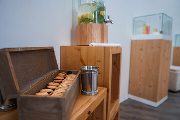 Refreshment area featuring cookies, infused water, and candies. Clean, inviting setting with wooden furniture and light walls. Ideal for office events or waiting rooms.
