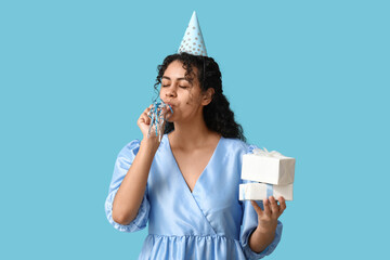 Beautiful young African-American woman in party hat with whistle and gift boxes on blue background