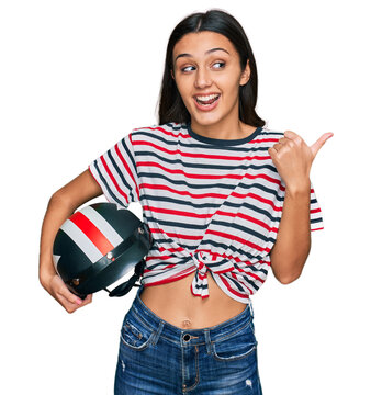 Young hispanic girl holding motorcycle helmet pointing thumb up to the side smiling happy with open mouth