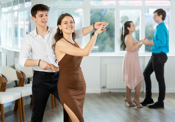 Stylish emotional young hispanic guy in white shirt and black trousers enjoying impassioned merengue with female partner wearing elegant dress in latin dance class. Amateur dancing concept - obrazy, fototapety, plakaty