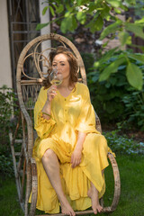 woman relaxing in the garden with a glass of wine. woman in a yellow dress sitting in a wicker chair in the garden with a glass - 780927822