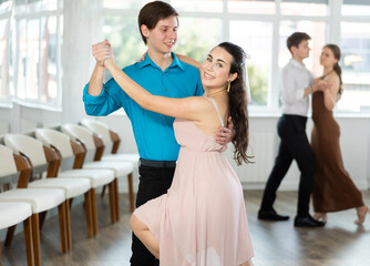 Smiling young brunette in light flowing dress practicing sensual bachata with guy in dance class....