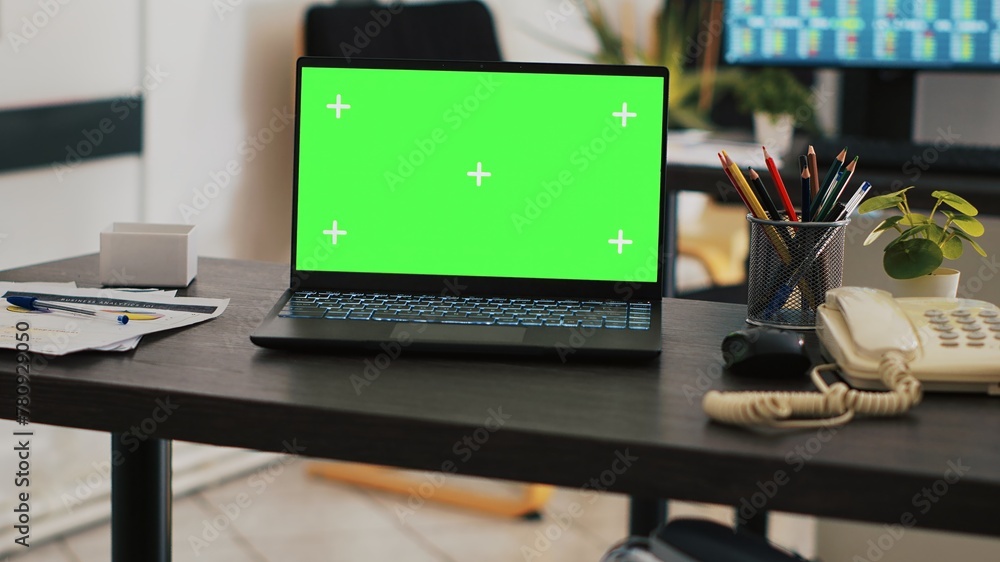 Poster focus on green screen laptop on office desk with computer in blurry background showing stock exchang