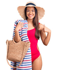 Young beautiful latin girl wearing swimwear and summer hat holding beach towel and bag pointing...