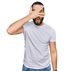 Young man with beard wearing casual white t shirt peeking in shock covering face and eyes with hand, looking through fingers with embarrassed expression.