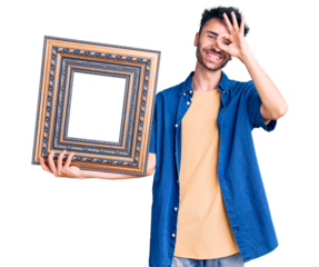 Young hispanic man holding empty frame smiling happy doing ok sign with hand on eye looking through fingers