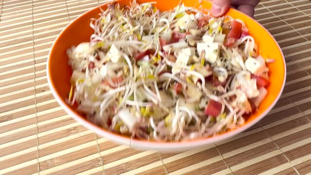 A fresh vegetable salad. Tomatoes, onions, apples, mound of sprouted mung beans in orange bowl on bamboo background. Chef's hand removes orange bowl from the table. Close up.