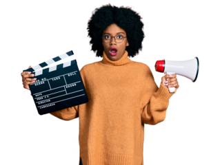 Young african american woman holding video film clapboard and megaphone in shock face, looking skeptical and sarcastic, surprised with open mouth