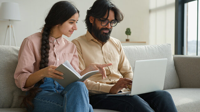 Diverse couple on couch with laptop pay bills loan payments planning household budget arabian woman wife read notes in notebook indian man husband typing data email on computer family business at home