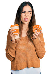 Beautiful hispanic woman eating doughnut and drinking coffee sticking tongue out happy with funny expression.