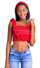 Young african american woman wearing casual summer clothes smiling with happy face looking and pointing to the side with thumb up.