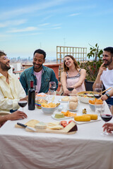 Vertical. Group of laughing young people enjoying lunch together outdoor. Gathering of cheerful friends celebrating meal party on terrace. Concept of millennial friendship and having fun on weekend