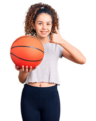 Beautiful kid girl with curly hair holding basketball ball smiling happy and positive, thumb up...