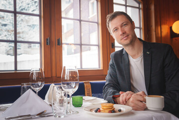 Man in dark blazer enjoying pancakes in cozy restaurant with snowy view. Warm ambiance, fine dining setup, relaxed smile, coffee cup, location unknown.