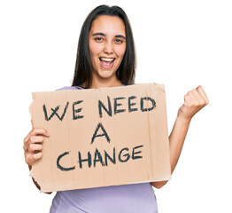 Young hispanic woman holding we need a change banner screaming proud, celebrating victory and success very excited with raised arms