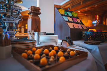 Buffet setup with fruits, plates, silverware, beverage dispenser, pepper grinder, snack packets display, all in a cozy, elegant wooden interior, focus on fruits. - obrazy, fototapety, plakaty
