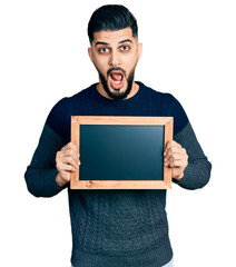 Young arab man with beard holding blackboard afraid and shocked with surprise and amazed expression, fear and excited face.