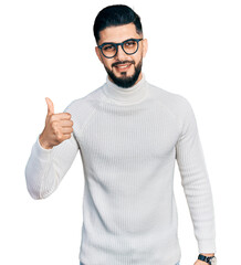 Naklejka premium Young arab man with beard wearing elegant turtleneck sweater and glasses doing happy thumbs up gesture with hand. approving expression looking at the camera showing success.