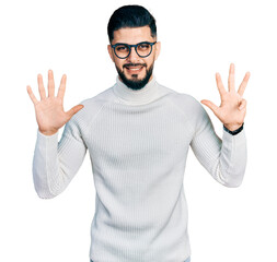 Naklejka premium Young arab man with beard wearing elegant turtleneck sweater and glasses showing and pointing up with fingers number nine while smiling confident and happy.