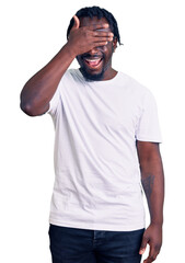 Fototapeta premium Young african american man with braids wearing casual white tshirt smiling and laughing with hand on face covering eyes for surprise. blind concept.