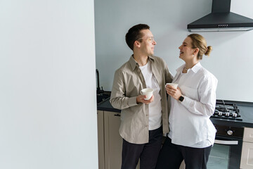 A couple shares a warm, convivial moment in their sleek kitchen, both clad in neutral tones, with mugs in hand, exuding a casual, comfortable domesticity. - obrazy, fototapety, plakaty