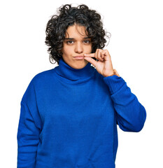 Young hispanic woman with curly hair wearing turtleneck sweater mouth and lips shut as zip with...