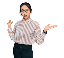 Young hispanic girl wearing casual clothes and glasses clueless and confused expression with arms...