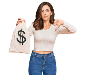 Young brunette woman holding dollars bag with angry face, negative sign showing dislike with thumbs...