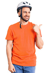 Naklejka premium Young handsome man wearing bike helmet smiling with happy face looking and pointing to the side with thumb up.