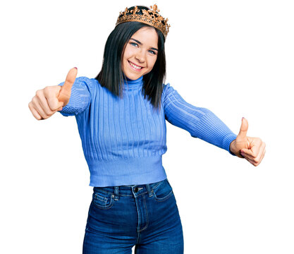 Young brunette woman with blue eyes wearing princess crown approving doing positive gesture with hand, thumbs up smiling and happy for success. winner gesture.