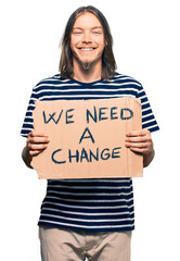 Handsome caucasian man with long hair holding we need a change banner looking positive and happy...