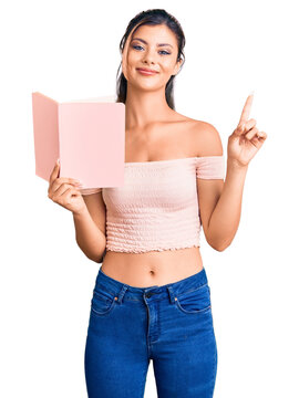 Young beautiful woman holding book surprised with an idea or question pointing finger with happy face, number one