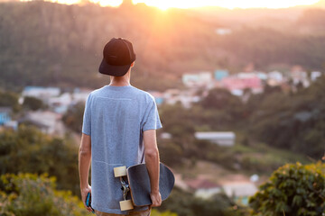 Man standing at the top of a hill, holding his longboard, with a view of a small town in a valley...